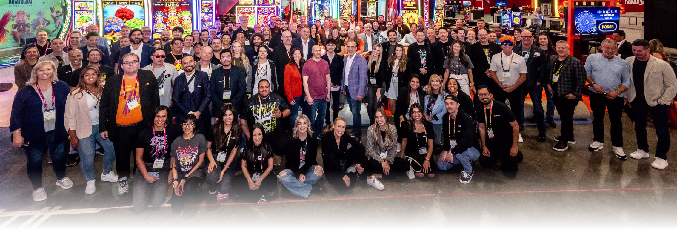 Large group of diverse people posing together indoors at a gaming or technology event with slot machines and electronic displays in the background.