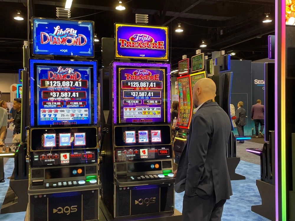 Man in suit standing and playing two illuminated slot machines named Liberty Diamond and Triple Treasure in a casino or gaming exhibition hall.