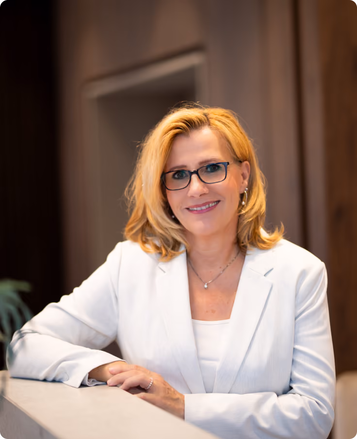 Frau mit blonden Haaren und Brille lächelt und lehnt an einer hellen Arbeitsfläche.