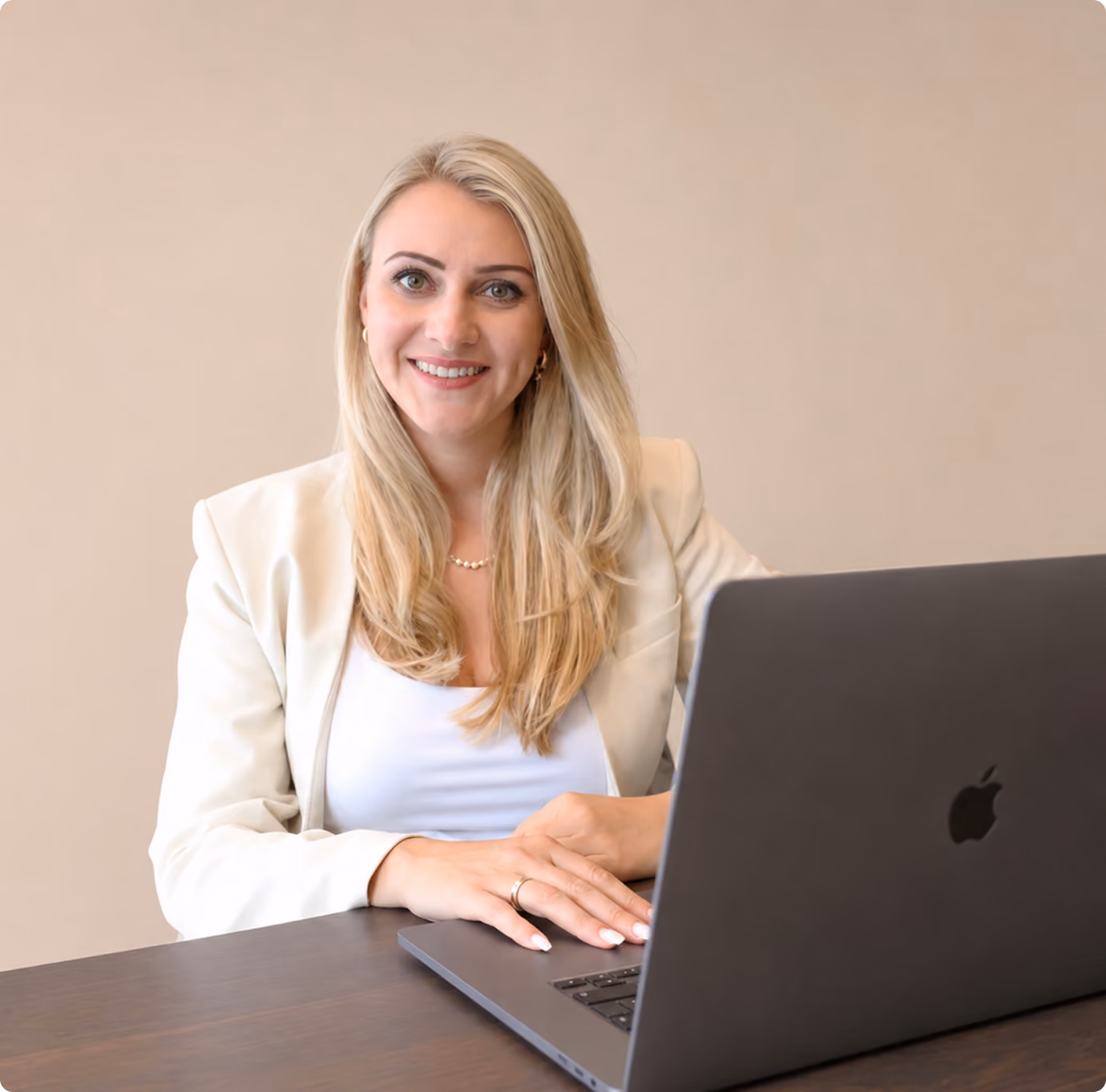 Lächelnde blonde Frau in weißem Blazer sitzt an Tisch mit offenem Laptop.