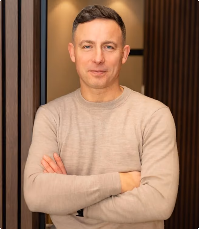 Mann mit kurzen braunen Haaren und beige Pullover steht mit verschränkten Armen vor dunklem Hintergrund.