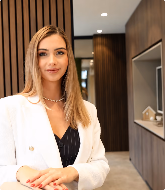 Junge Frau mit blondem Haar und weißem Blazer lächelt in modernem Büro mit Holzvertäfelung.