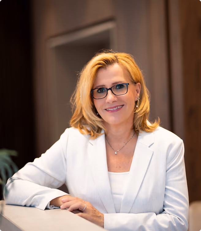 Blonde Frau mit Brille und weißem Blazer lehnt lächelnd an einem Tisch.