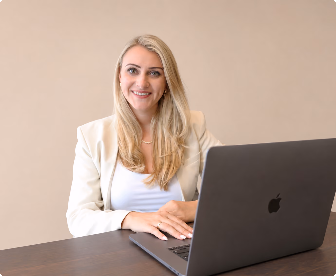 Frau mit langen blonden Haaren lächelt und sitzt am Tisch mit einem aufgeklappten grauen Laptop.