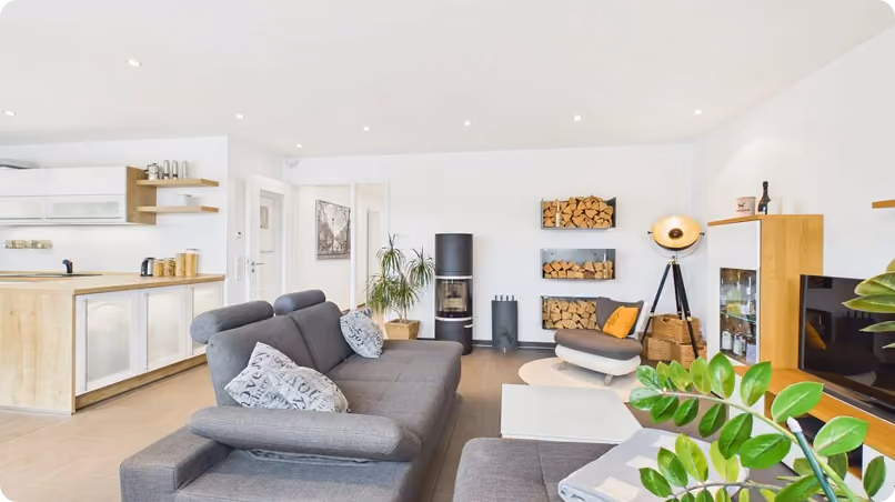Modern Wohnzimmer mit grauer Couch, Holzofen, Kaminholzregalen und offener Küche im Hintergrund.