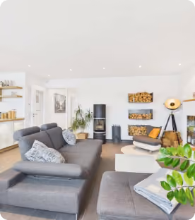 Modern Wohnzimmer mit grauem Sofa, Holzofen, Pflanzen und Bücherregalen.
