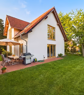 Modern white house with red roof, patio furniture, grill, and green lawn under blue sky.