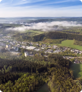 Luftaufnahme einer Stadt mit Wald- und Wiesenflächen unter blauem Himmel mit Wolken.