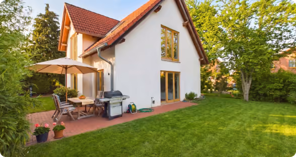 Haus mit rotem Dach, Terrasse mit Sonnenschirm, Grill und Garten mit grünem Rasen.