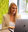 Junge Frau mit blondem Haar arbeitet an einem Laptop in einem hellen Raum mit grüner Wand.