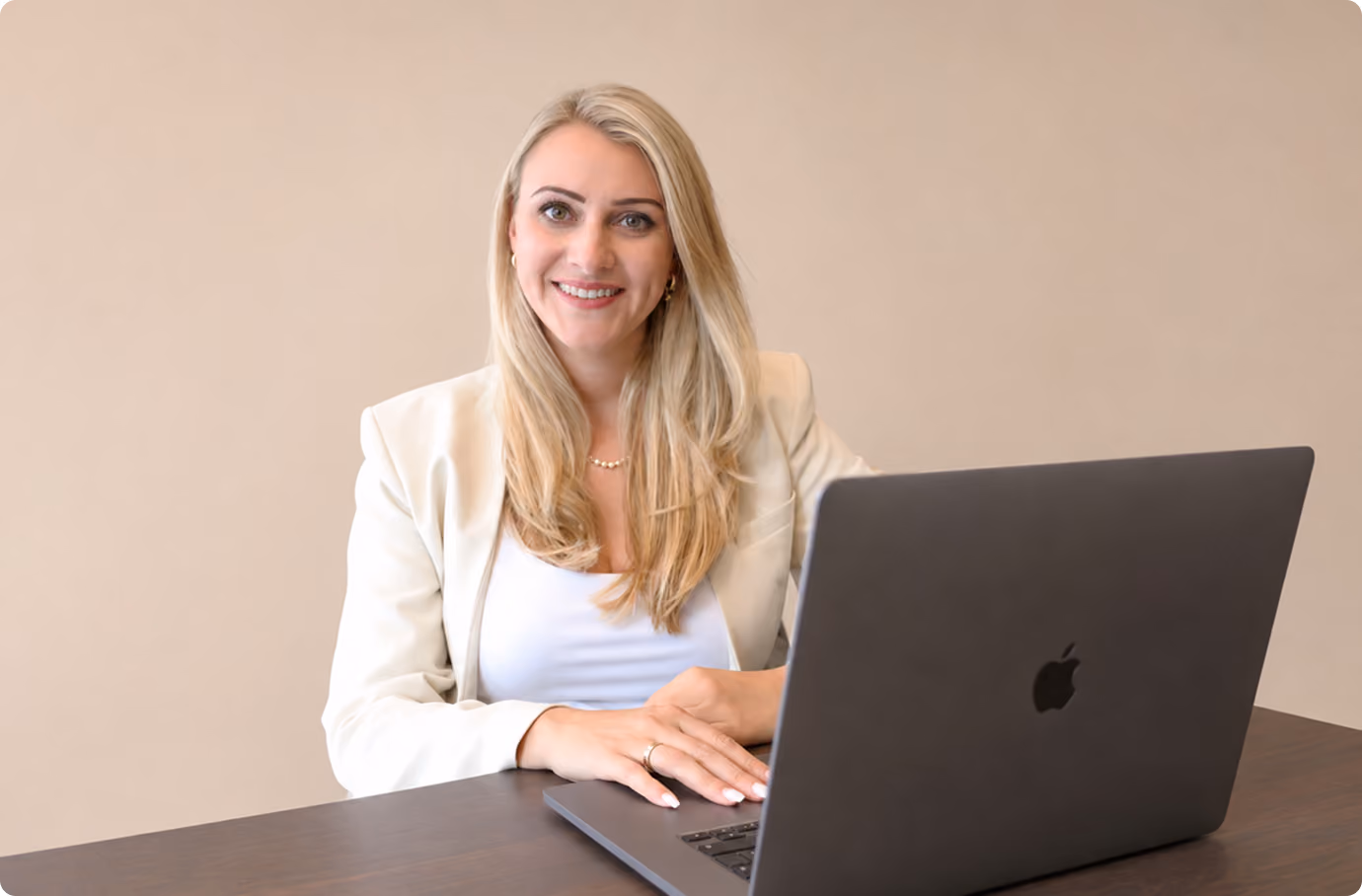 Frau mit langen blondem Haar lächelt am Tisch vor geöffnetem grauem Laptop.