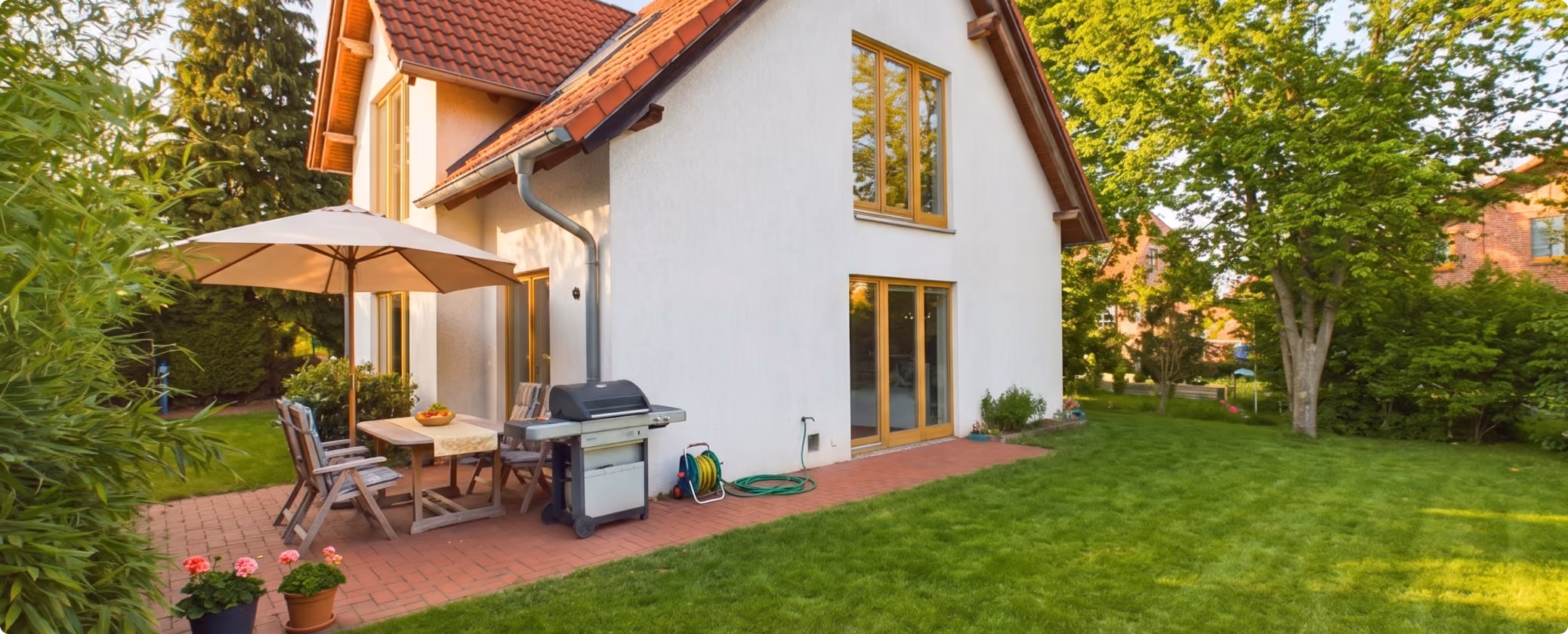 Haus mit rotem Ziegeldach, Terrasse mit Gartentisch, Stühlen, Sonnenschirm und Grill im Garten.