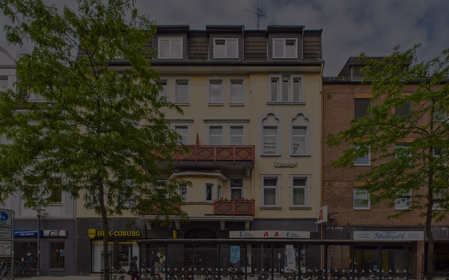 Straßenfront mit Baum, mehrstöckigen Gebäuden und Geschäften wie HUK-COBURG und Apotheke.