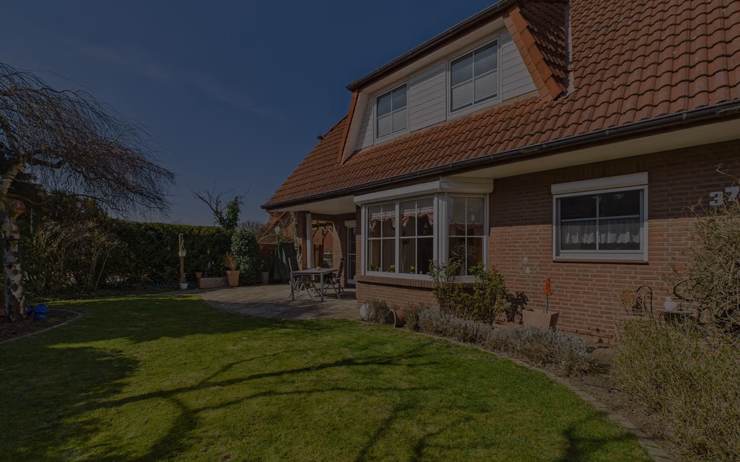 Ein Haus mit rotem Ziegeldach, Vorgarten mit grünem Rasen und Terrasse mit Tisch und Stühlen.