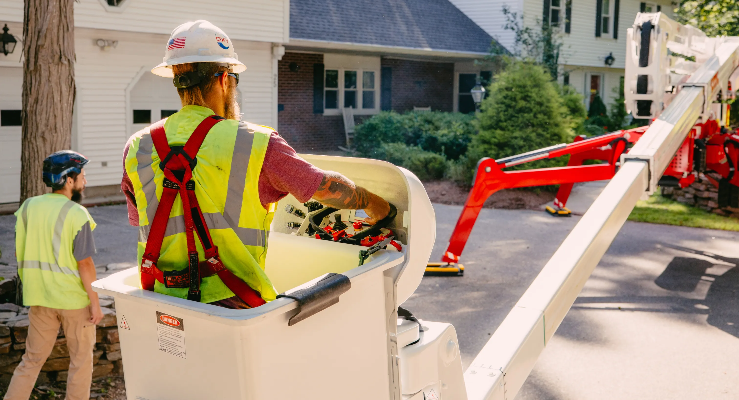 Worker in a safety harness operating a cherry picker lift near a residential house with another worker nearby.