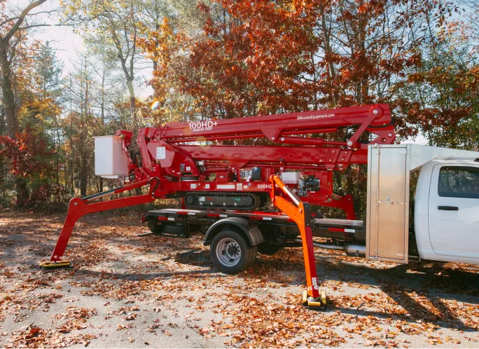 Red 100HD+ aerial lift mounted on a white truck with stabilizing legs extended on autumn leaves.