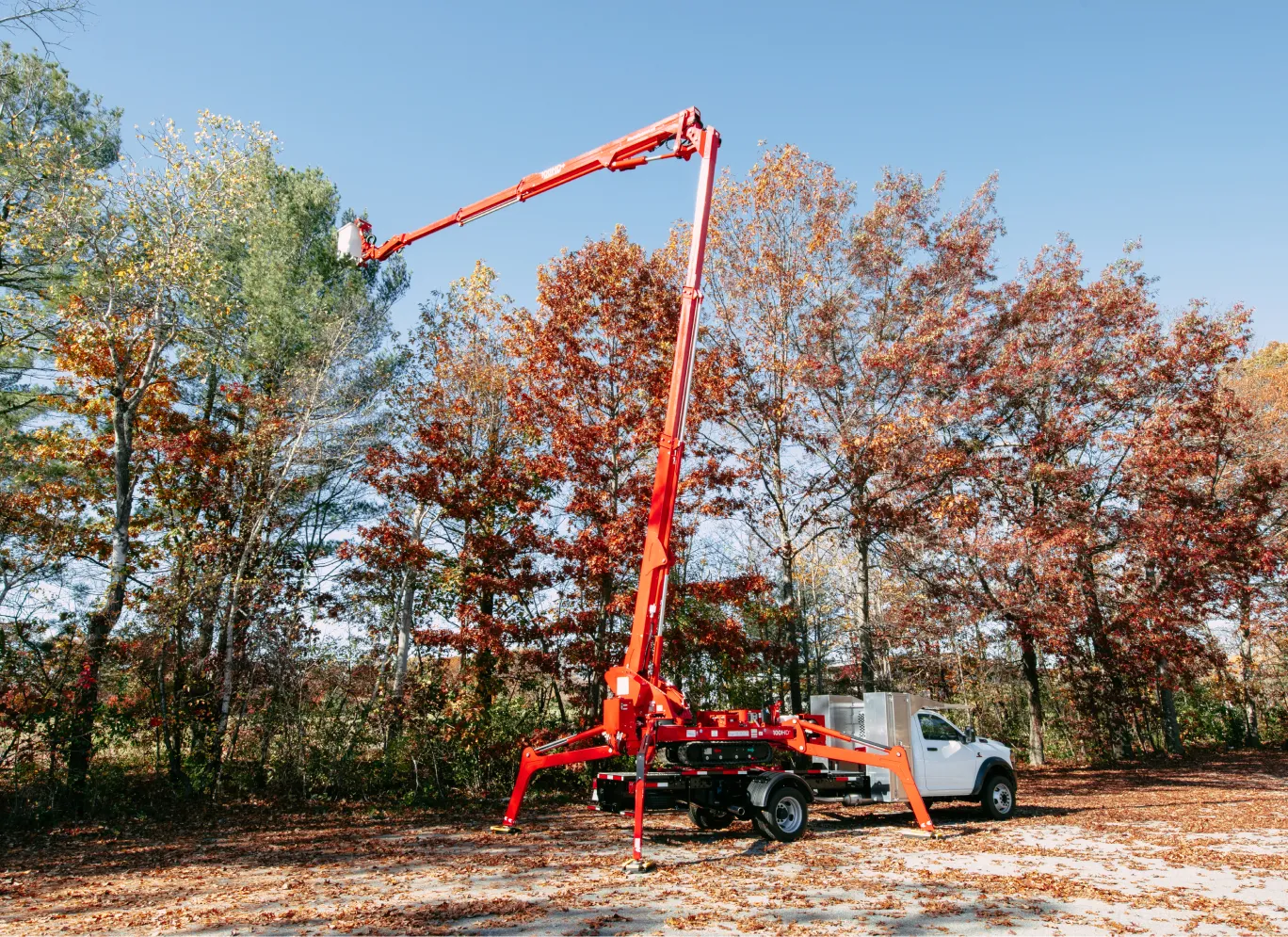 Red boom lift extended above fall trees with orange leaves beside white utility truck.