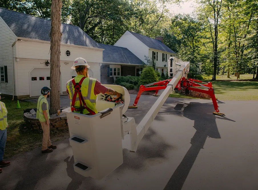 Worker in a spider lift bucket operating crane controls near a house driveway