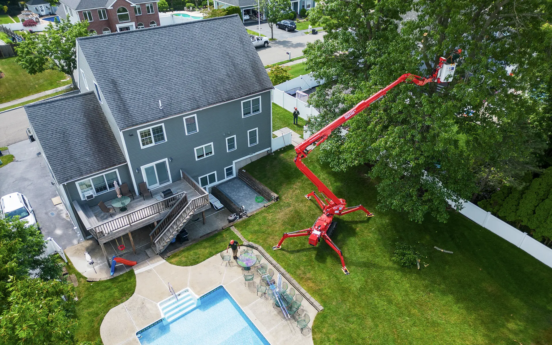 Workers using red hydraulic lift to trim tall tree next to suburban house with a backyard pool.