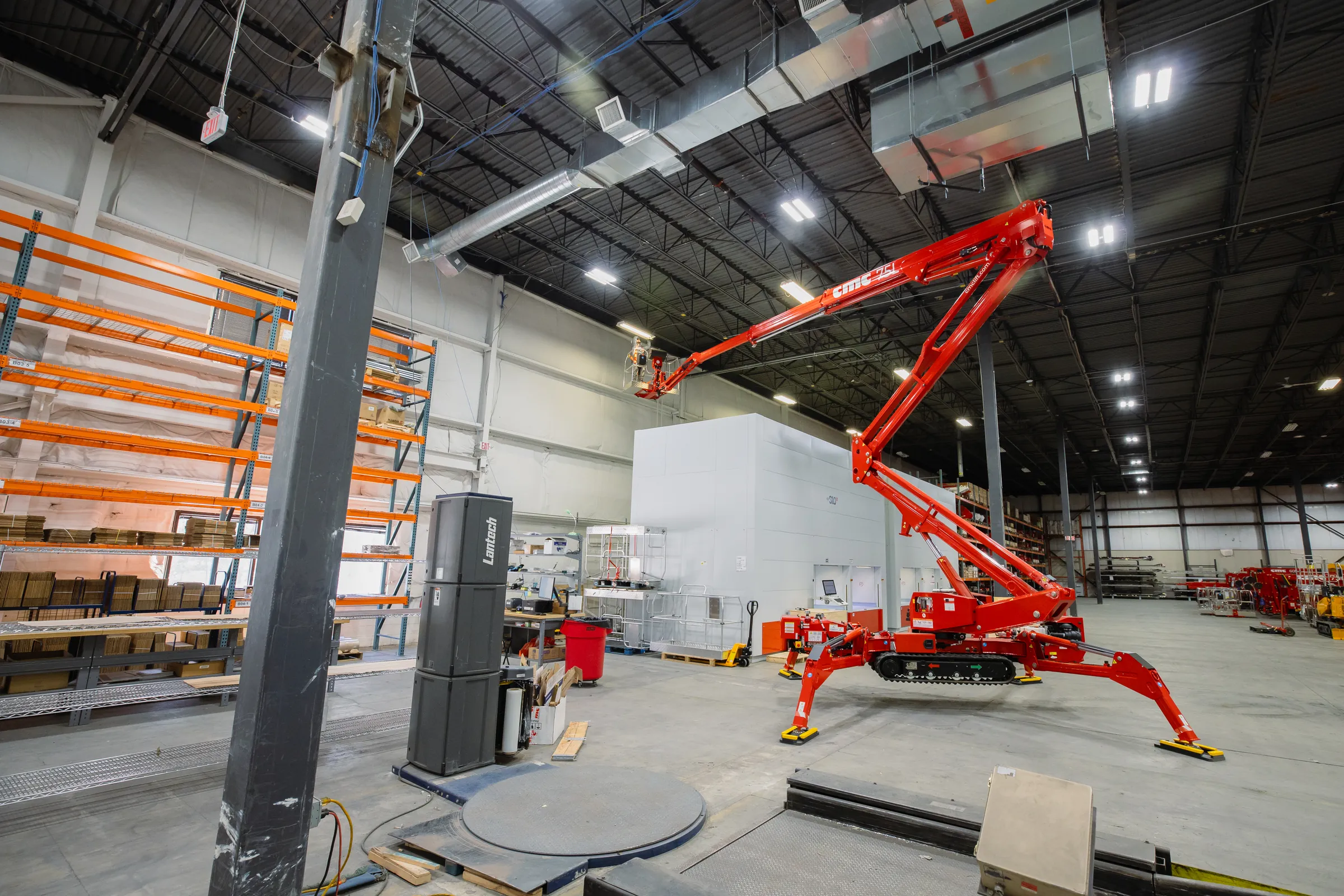 Red spider lift with extended arm inside a large industrial warehouse with shelves and equipment.