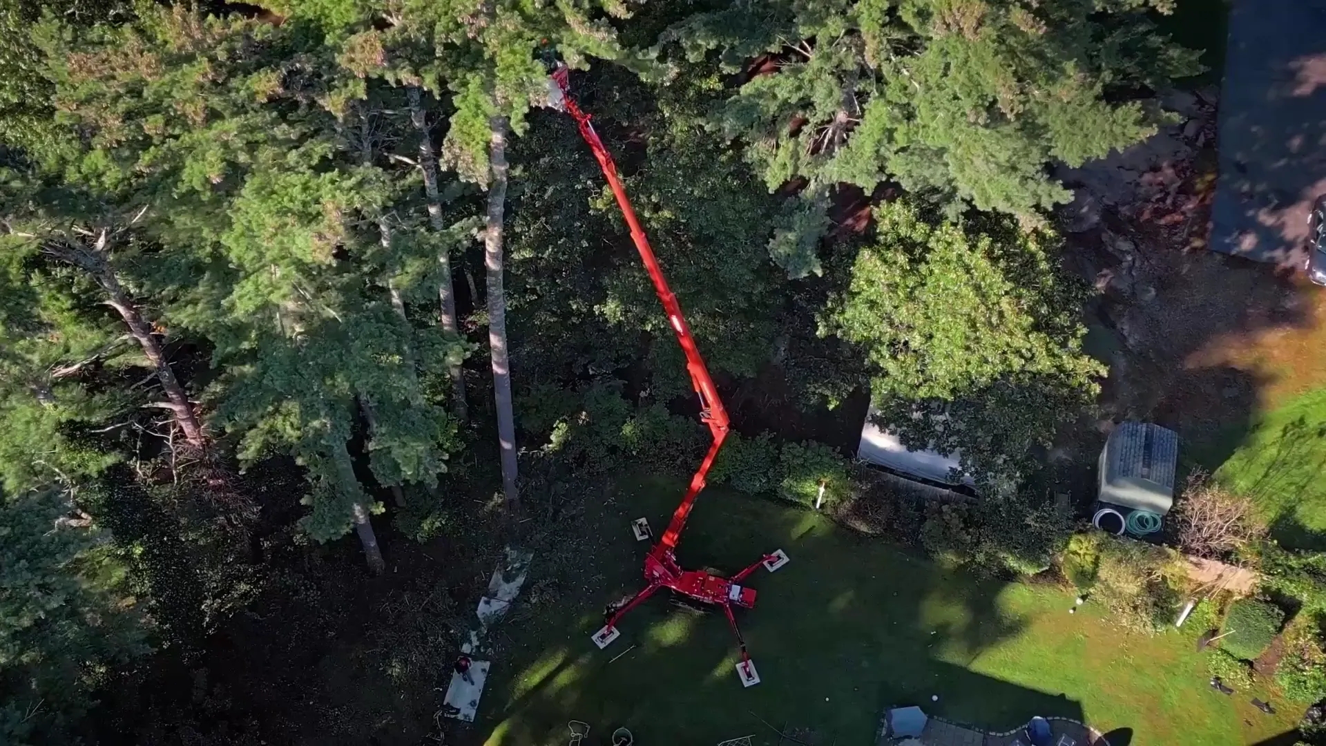 Aerial view of a red crane-like machine with extended arms on a lawn trimming tall trees around a house.