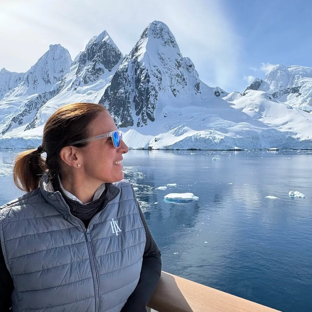 Woman wearing sunglasses looks at snowy mountains and ice floating on calm water under blue sky.