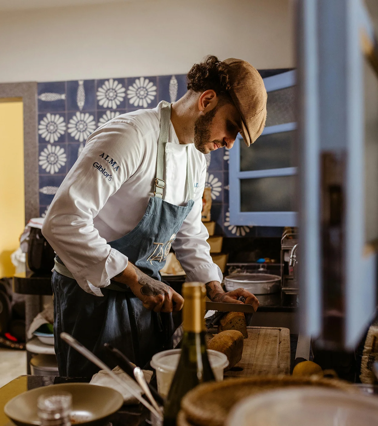 Chef in a white coat and apron cutting bread on a wooden board in a kitchen.
