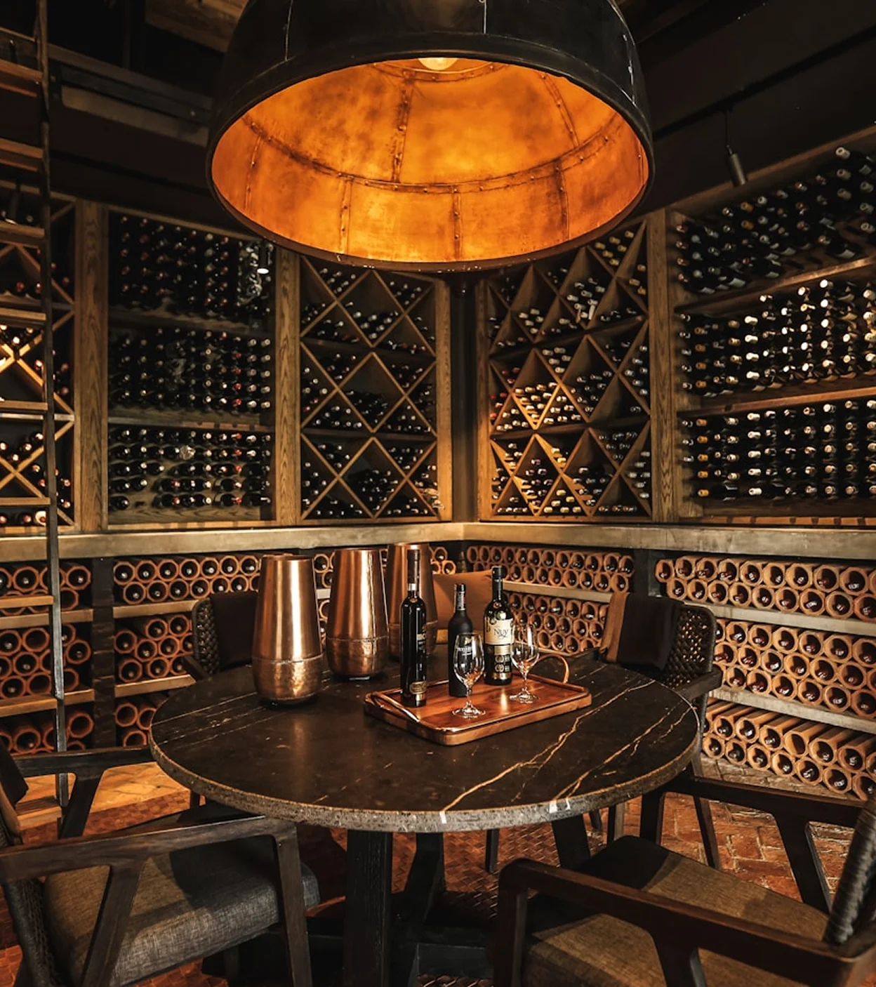 Cozy wine cellar with round table, four chairs, wine bottles on racks, and warm overhead light.
