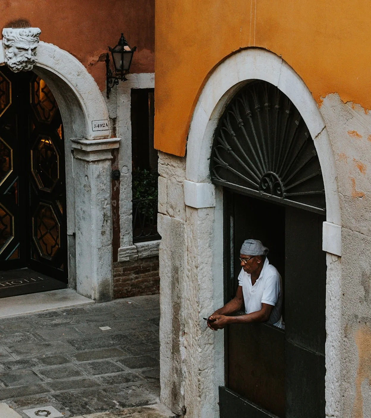 Man in white shirt and cap leaning on a half-door of an old yellow and stone building.