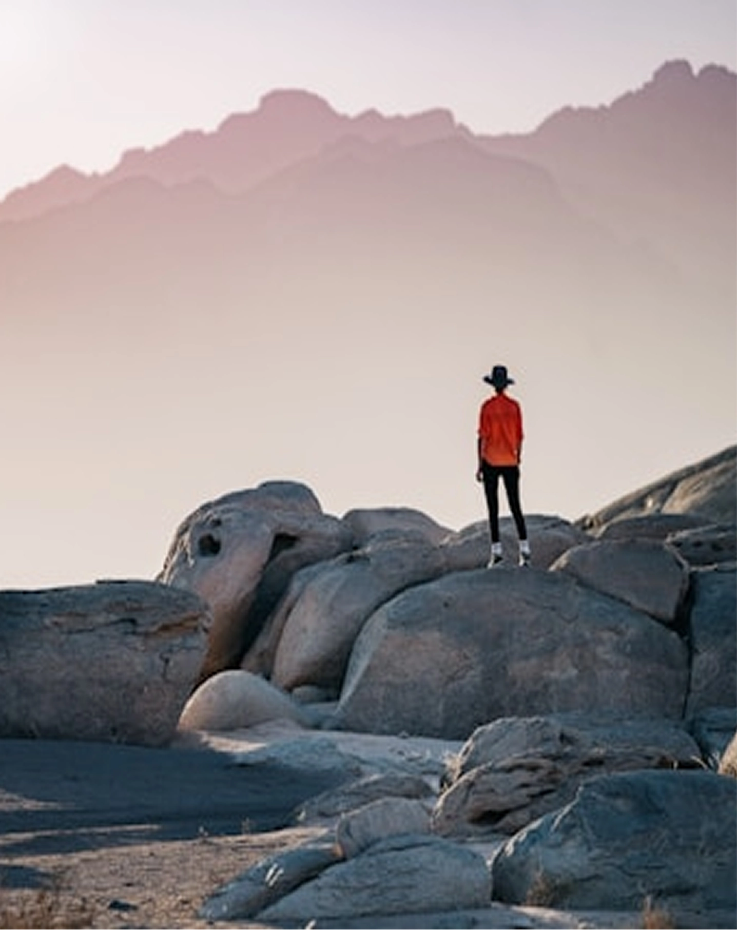 Person in an orange shirt and black hat standing on large rocks with mountains in the background.