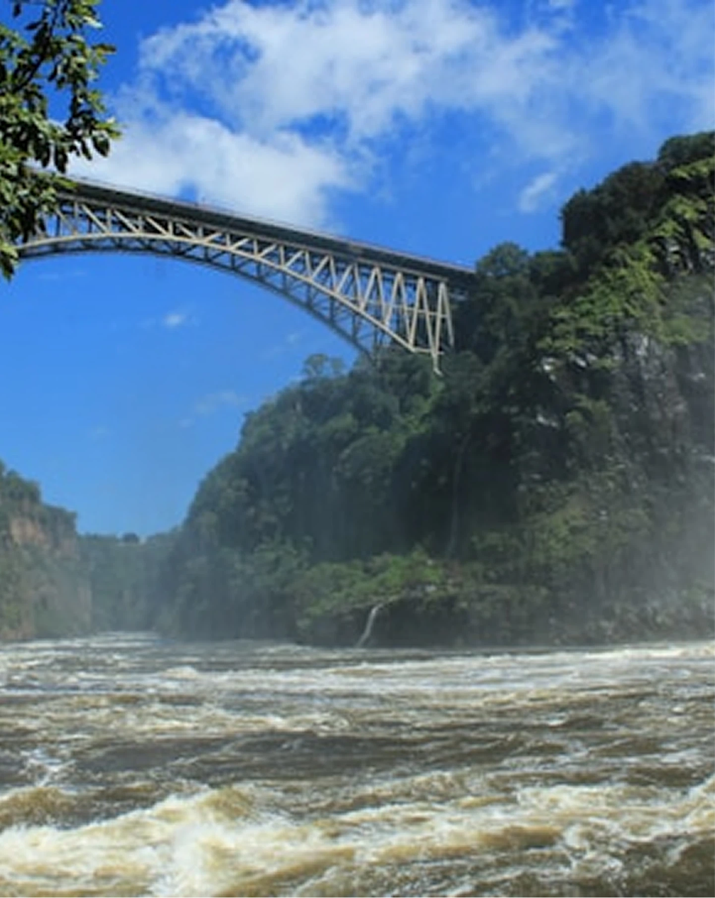 Metal arch bridge over turbulent river with forested cliffs under blue sky with clouds.