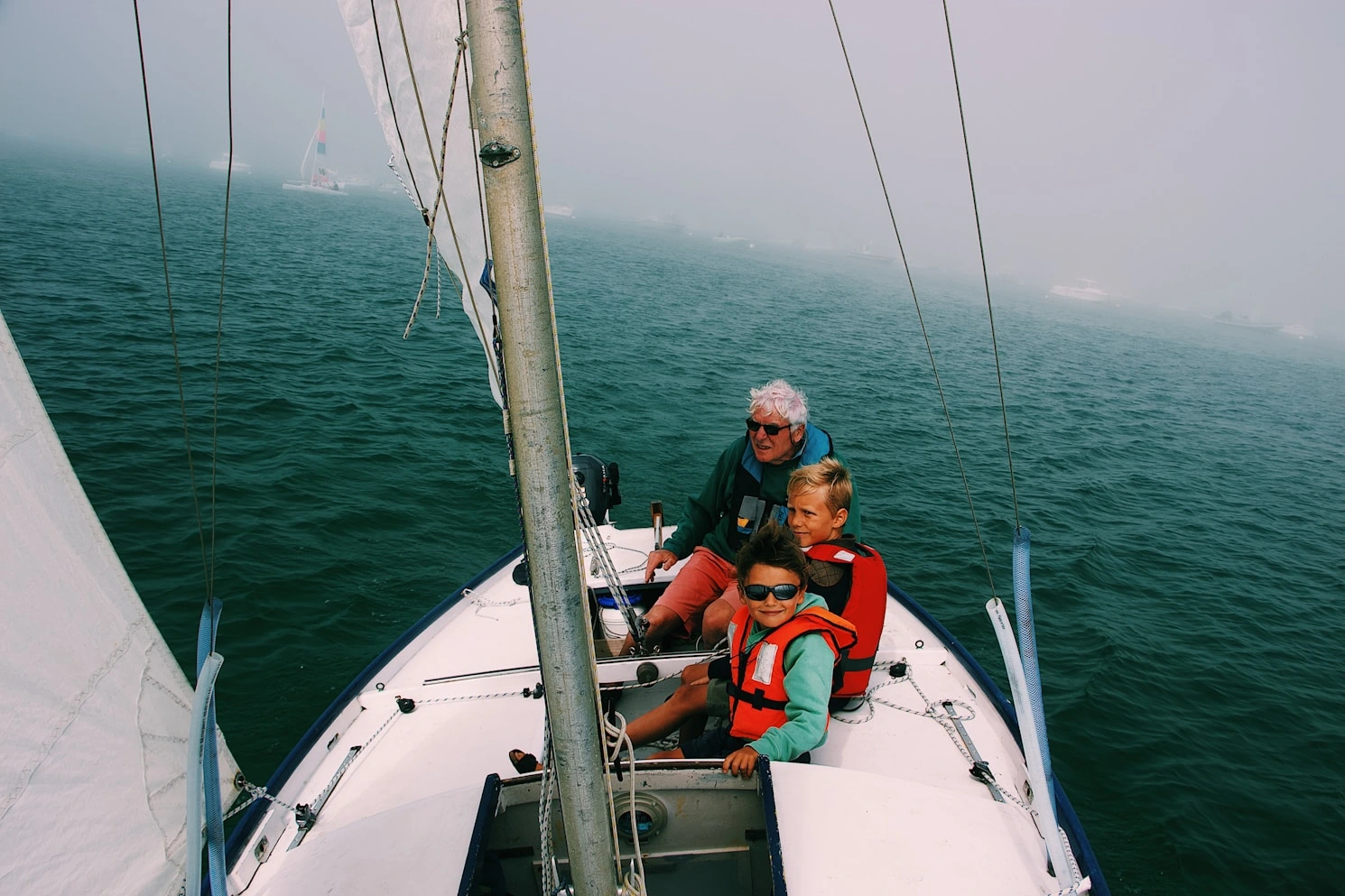 An older man and two boys wearing life jackets are sailing on a small boat on calm green water.