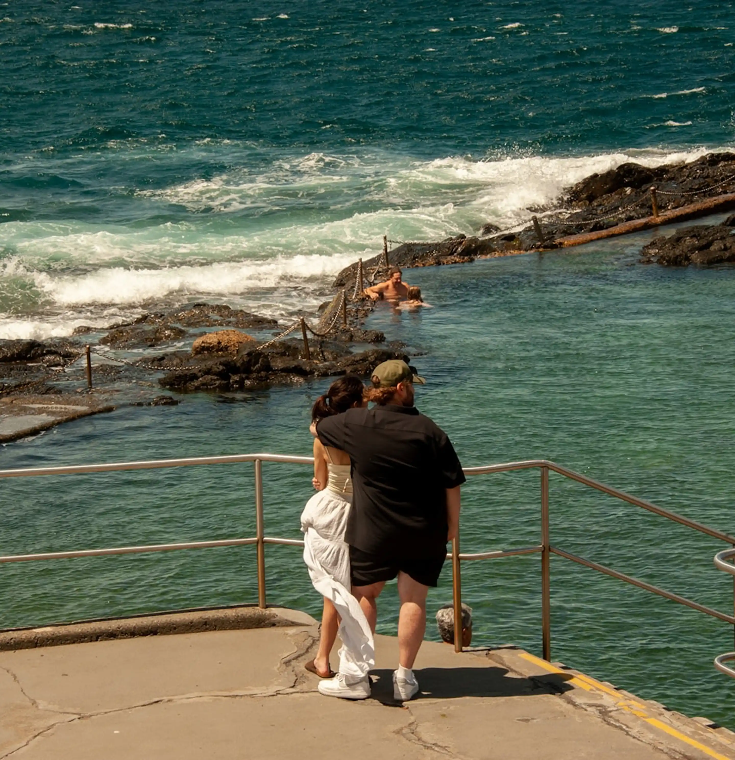 Couple with arm around each other stands by handrails looking toward natural rock pool and ocean waves.