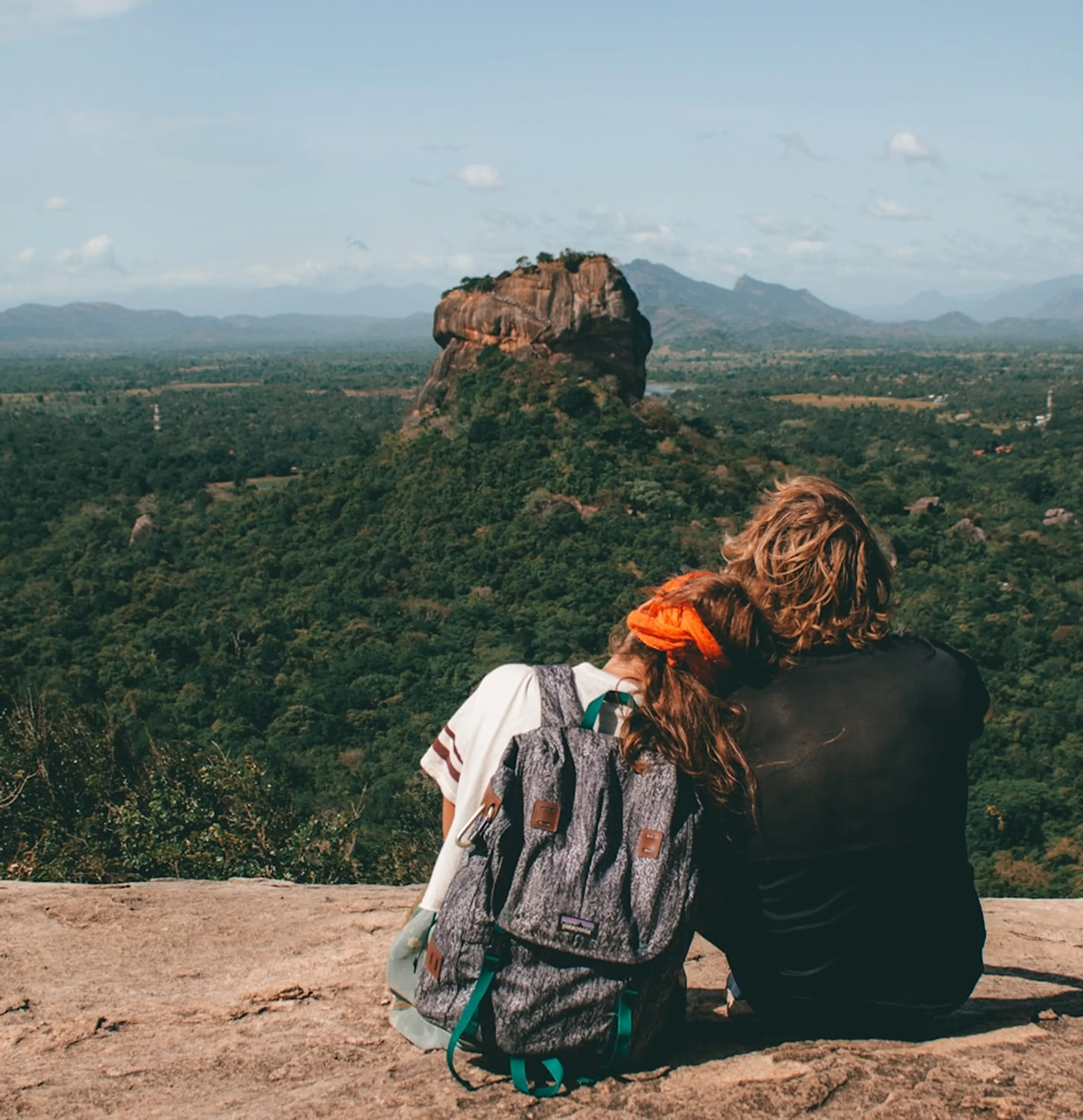 Two hikers sitting on a rock looking at a large forested rock formation surrounded by mountains.