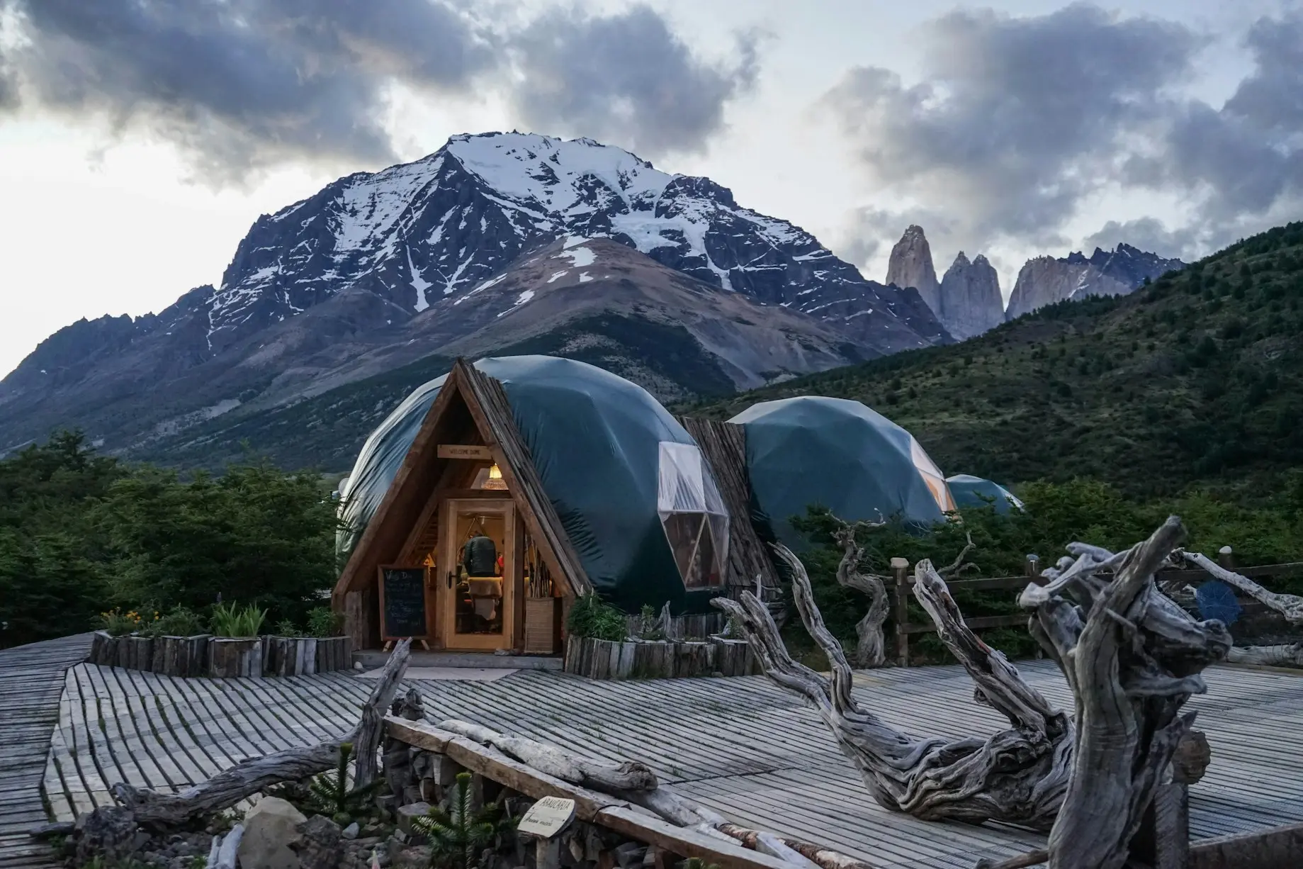Dome cabins with wooden frames near a snow-capped mountain under cloudy sky at dusk.