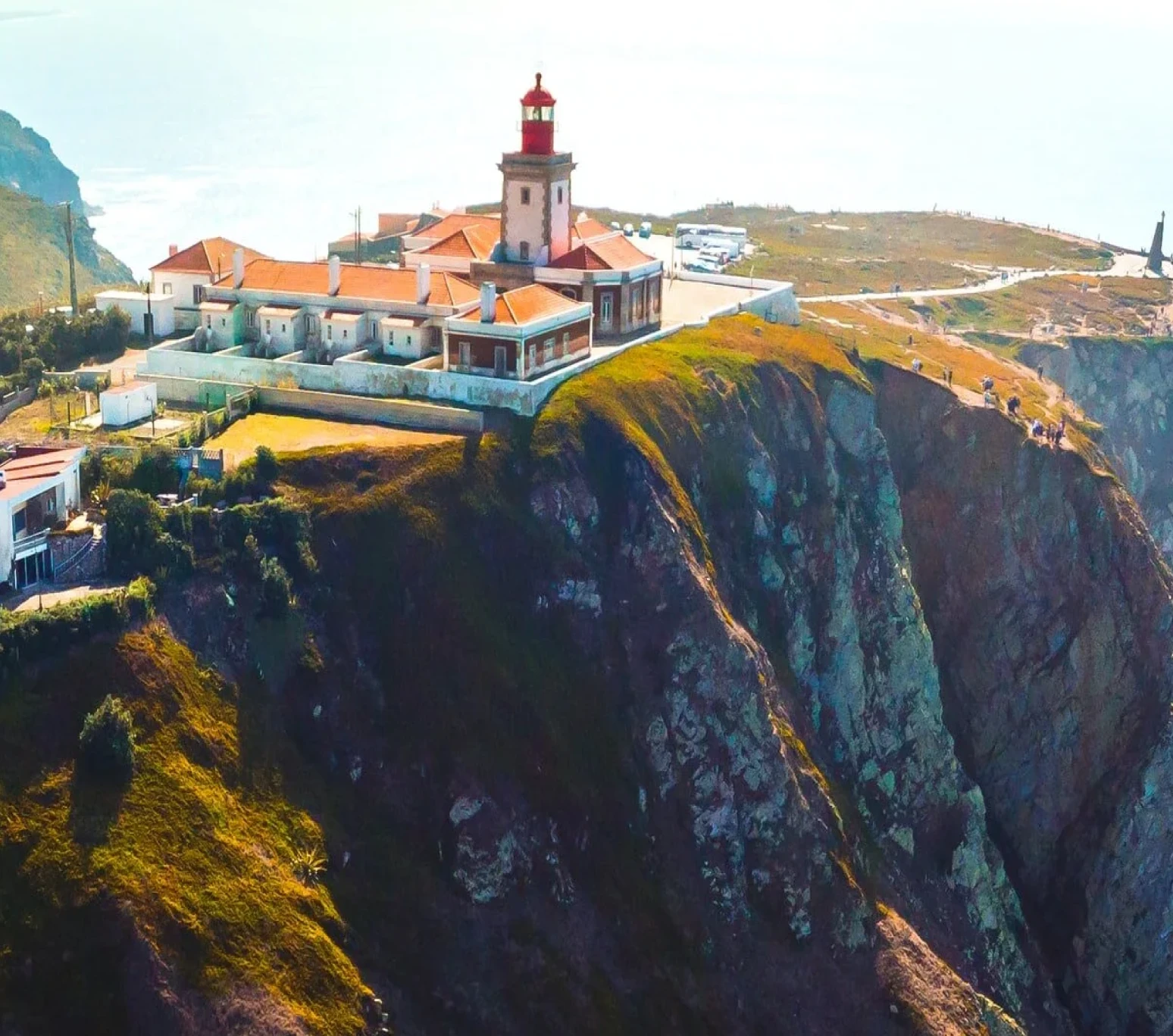 Lighthouse with red roof and tower on a green cliff overlooking the sea with visitors nearby.