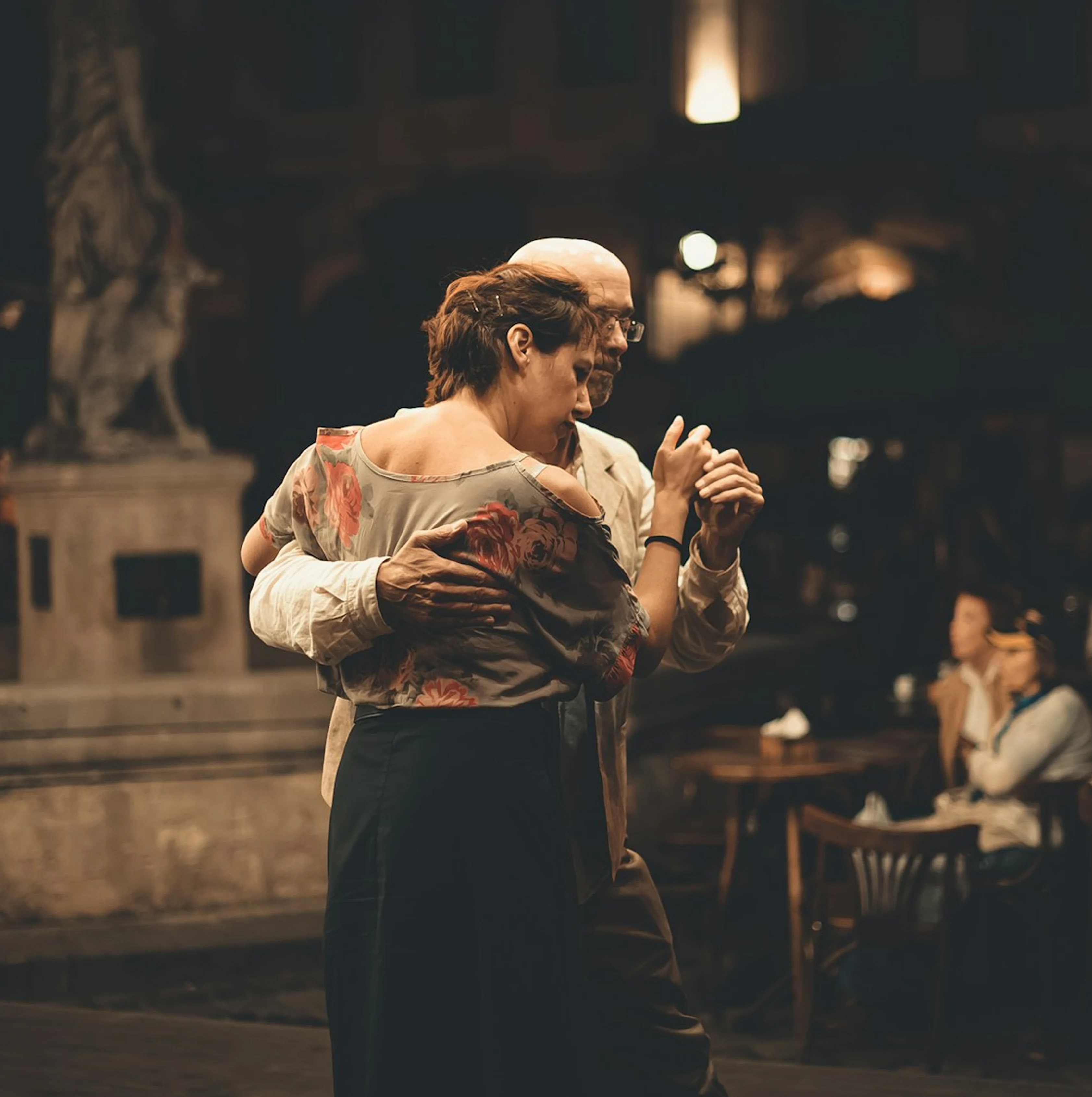 Couple dancing closely in warm lighting with background diners at an outdoor cafe.