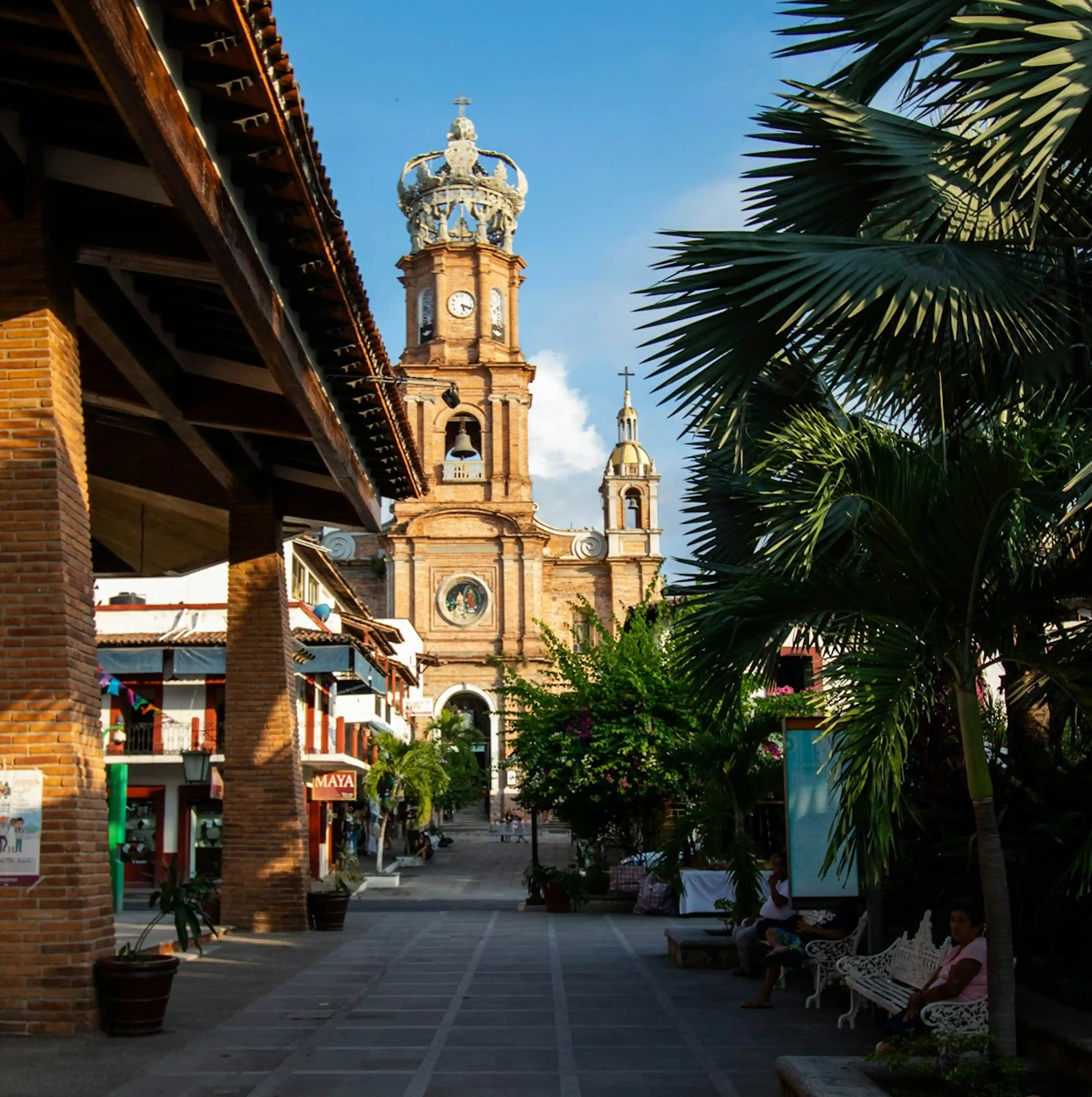 Church tower with crown top at end of plaza lined with palm trees and brick buildings