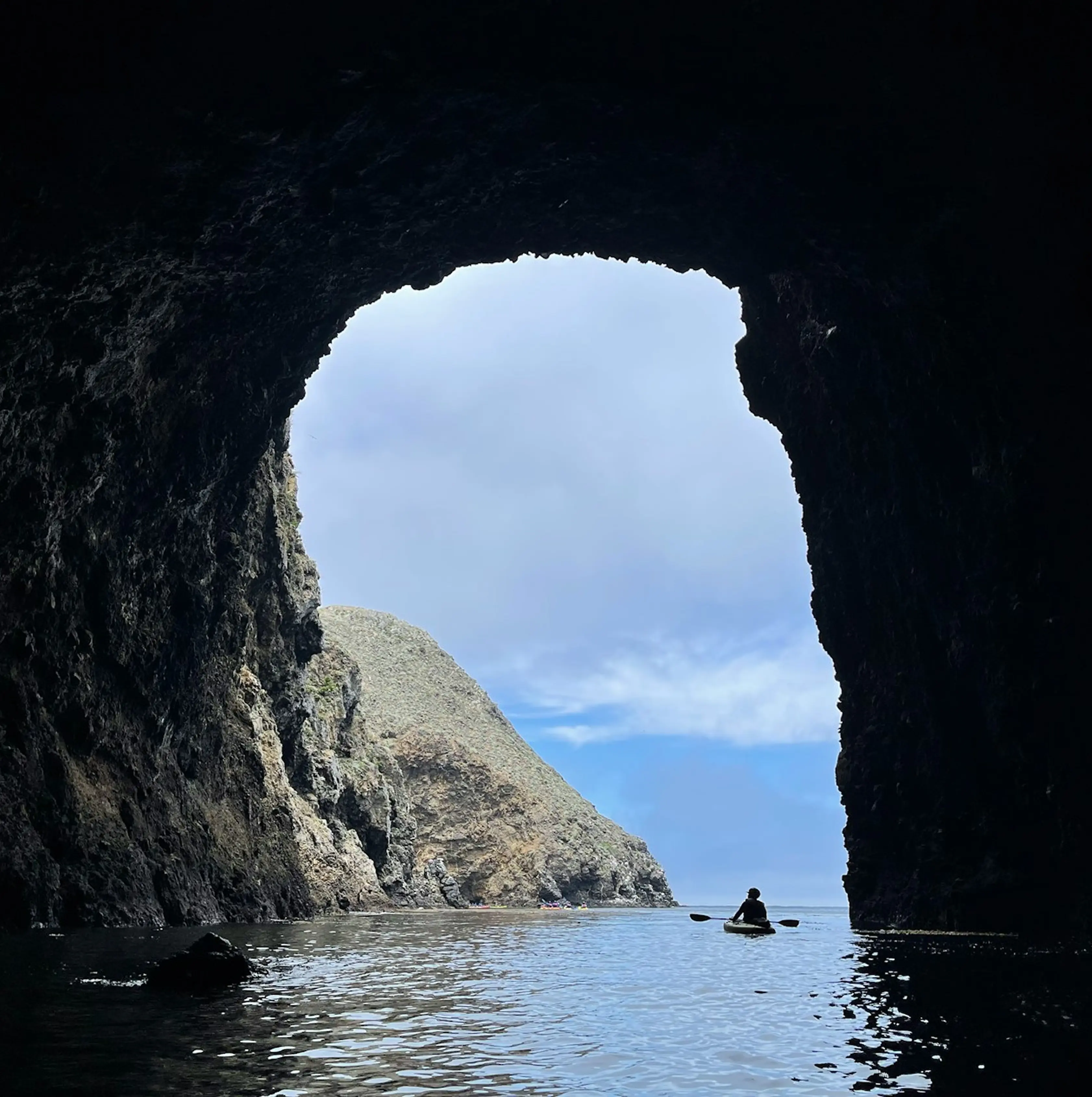 Person kayaking on water seen from inside a dark sea cave opening to rocky cliffs and sky.