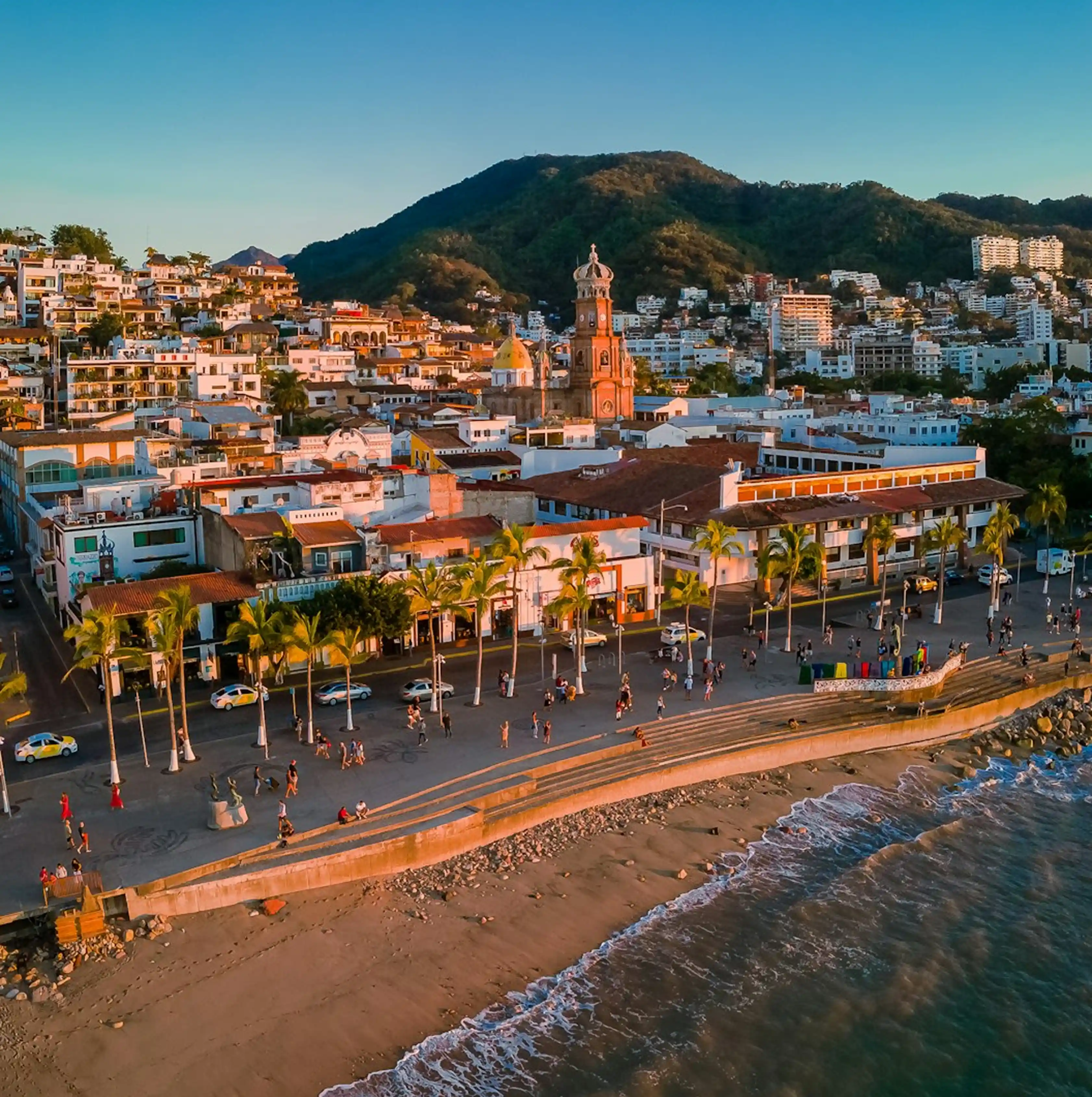 Coastal city at sunset with palm trees, beachfront promenade, and a church tower near green mountains.