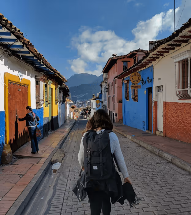 Person with backpack walking down a cobblestone street lined with colorful buildings under a cloudy sky.