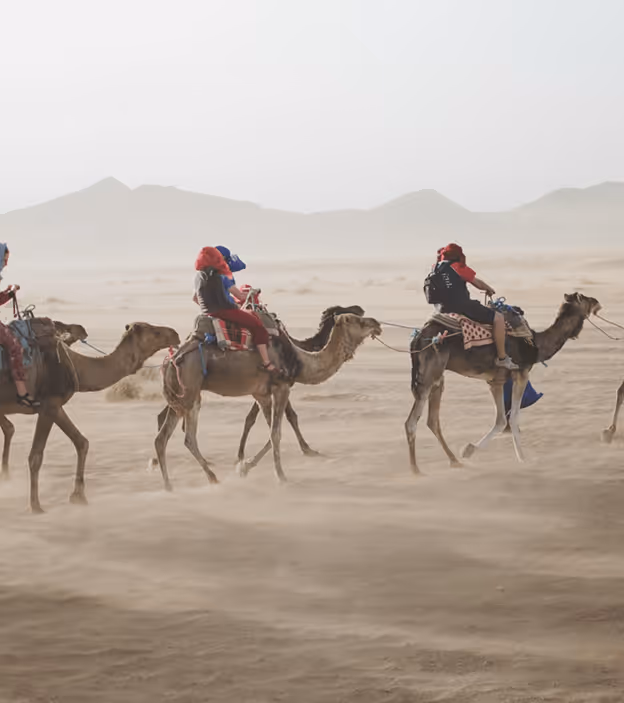 Group of people riding camels in a desert with mountains and dust in the background.