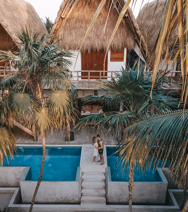 Couple standing on poolside stairs surrounded by palm trees in front of tropical thatched-roof huts.