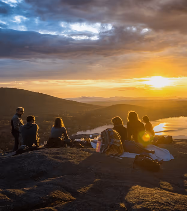 Group of people sitting on a rock at sunset overlooking hills and a reflective lake.