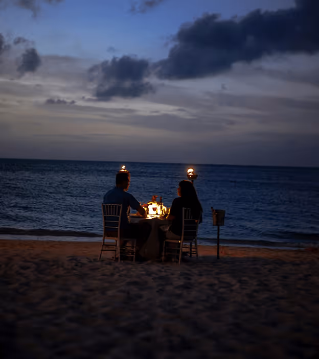 Couple having a candlelit dinner at night on a beach by the ocean with two torches.
