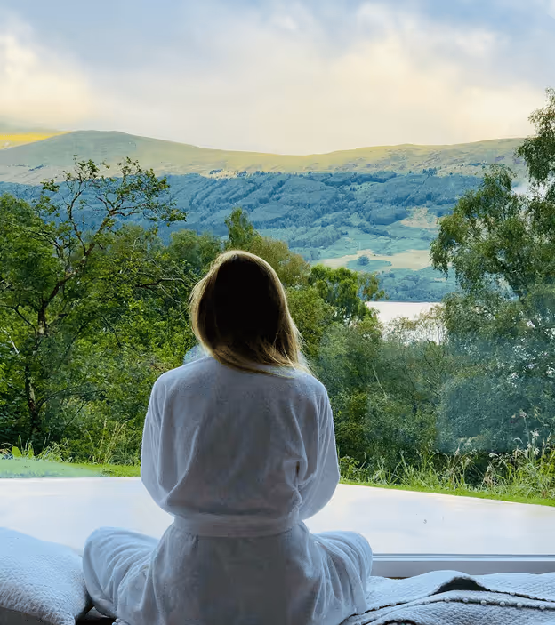 Person in white robe sitting by a window, looking at green hills and a lake in the distance.