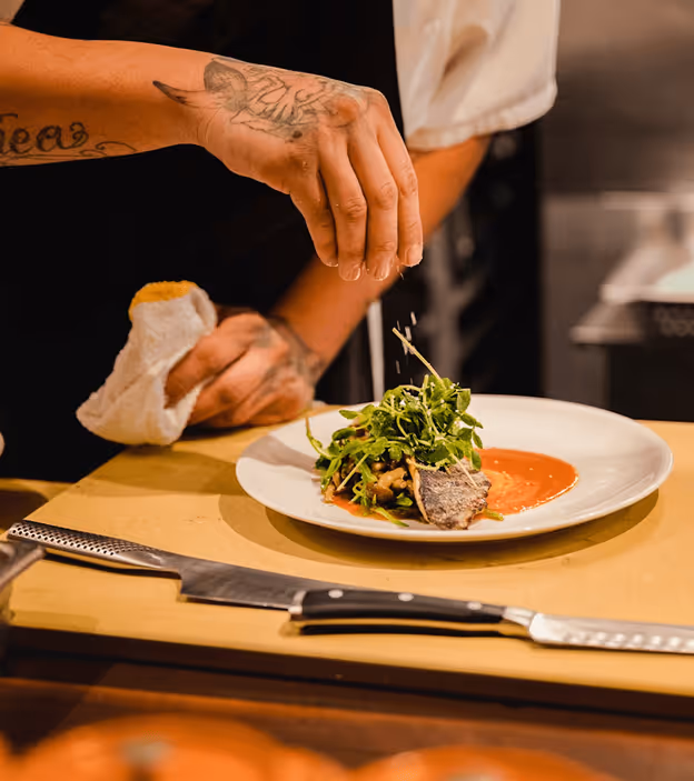 Chef with tattooed hand seasoning plated dish with greens and sauce in a kitchen setting.