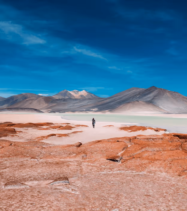 Person standing on reddish desert ground with a shallow lake and mountains under a blue sky.