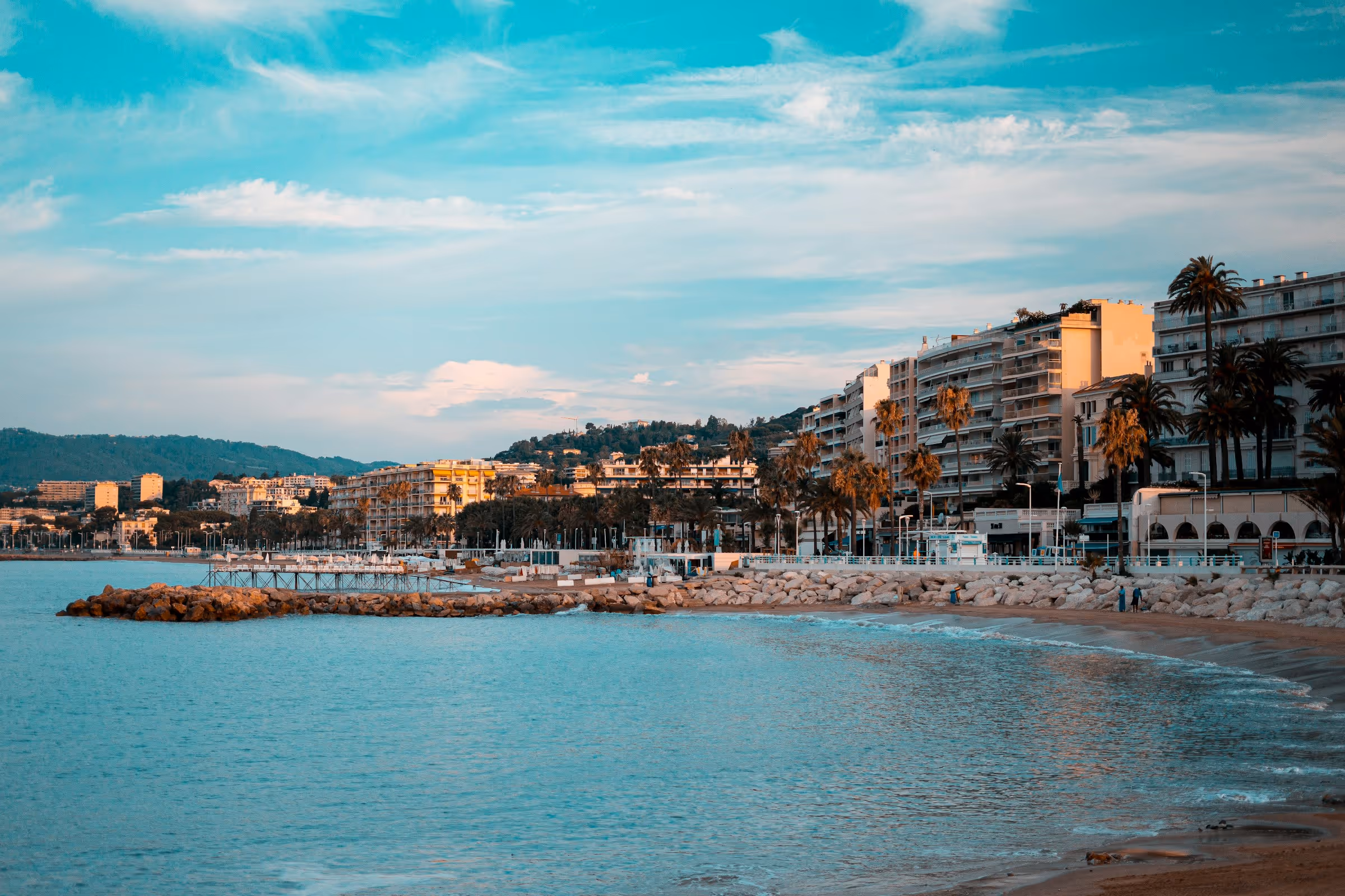 Coastal city shoreline with palm trees, modern buildings, mountains, and calm blue sea under a partly cloudy sky.