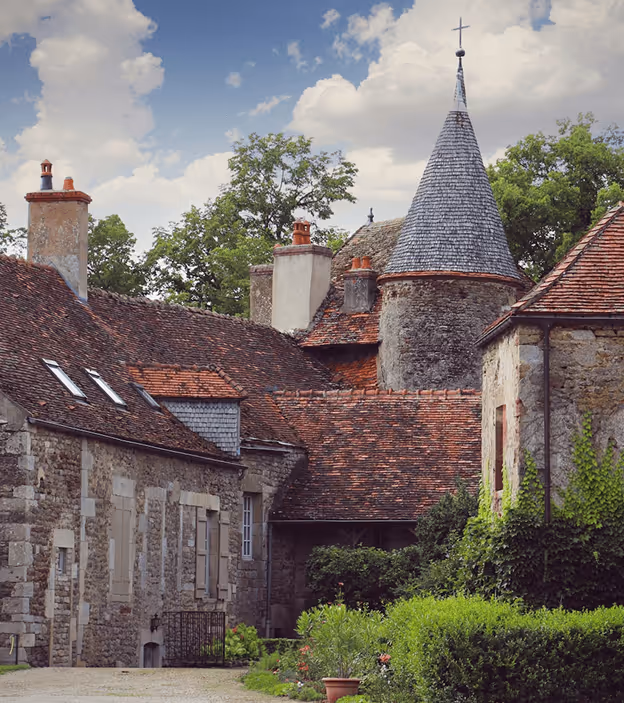 Stone buildings with red-tiled roofs and a pointed tower in a lush countryside garden.